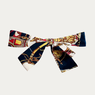 Une ceinture en tissu femme à imprimé baroque nouée autour d'un jean bleu sur un fond gris très clair uni